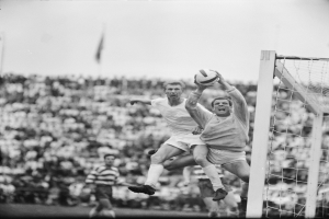 Zwei M├Ąnner beim F├╝├čballspielen vor einer Zuschauermenge, mit einem Torpfosten auf der rechten Seite und einer Flagge im Hintergrund, dargestellt in Schwarz-Wei├č.