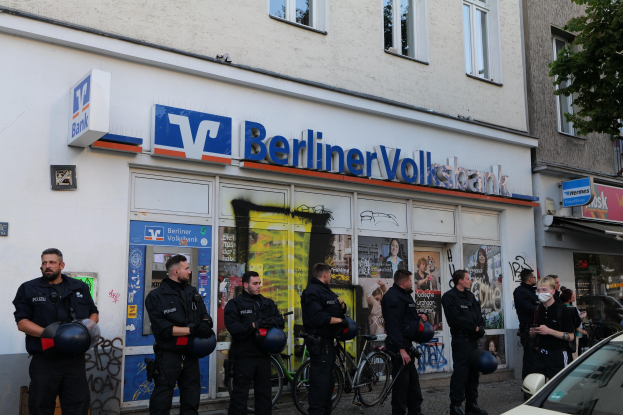 Eine Gruppe von Polizisten steht vor einer Berliner Volksbank, einige halten Helme, daneben ein geparktes Auto und ein Baum; im Hintergrund sind Gebäude mit Fenstern und Texttafeln zu sehen.