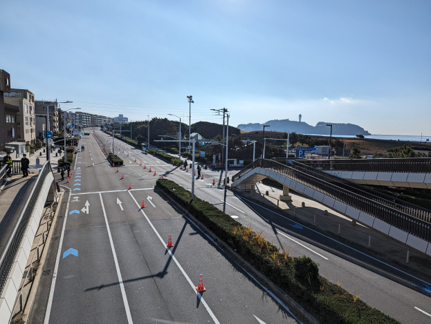 Straße mit Verkehrskegeln, eine Brücke mit Geländern, Laternenmasten, Gebäude mit Fenstern und einige Menschen; Hügel und bewölkter Himmel im Hintergrund, Teil des Takayama Bridge Projekts.