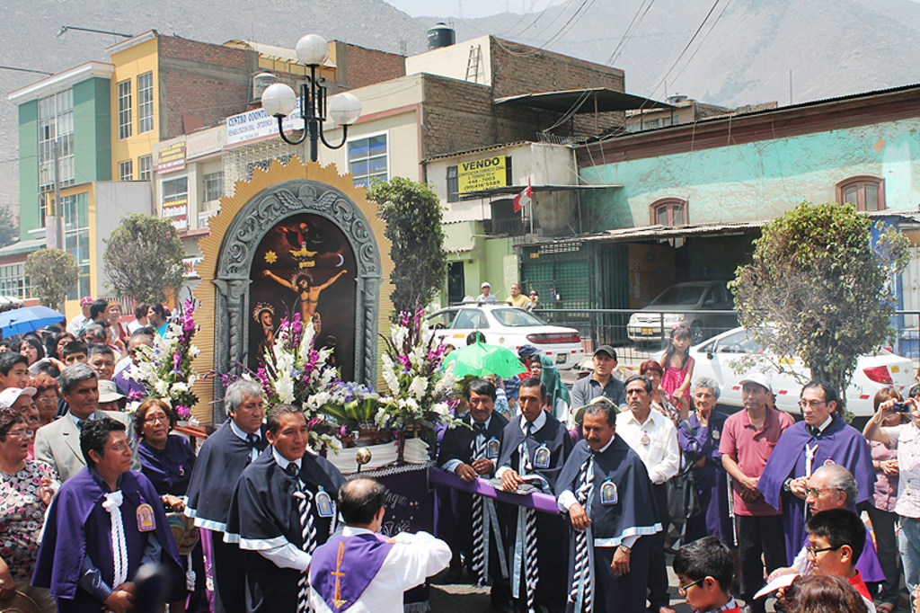 Menschen auf einer Straße vor der Stadt, mit einem Kreuz und Blumensträußen im Vordergrund, Autos im Hintergrund und Gebäude, Bäume und Straßenlaternen weiter hinten.