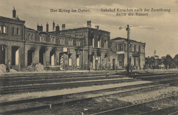 Schwarzes und weißes Foto des Bahnhofs Korschen in Ostern, Deutschland, mit Gleisen, Gebäuden, Strommasten, Menschen, Bäumen und Himmel im Hintergrund.