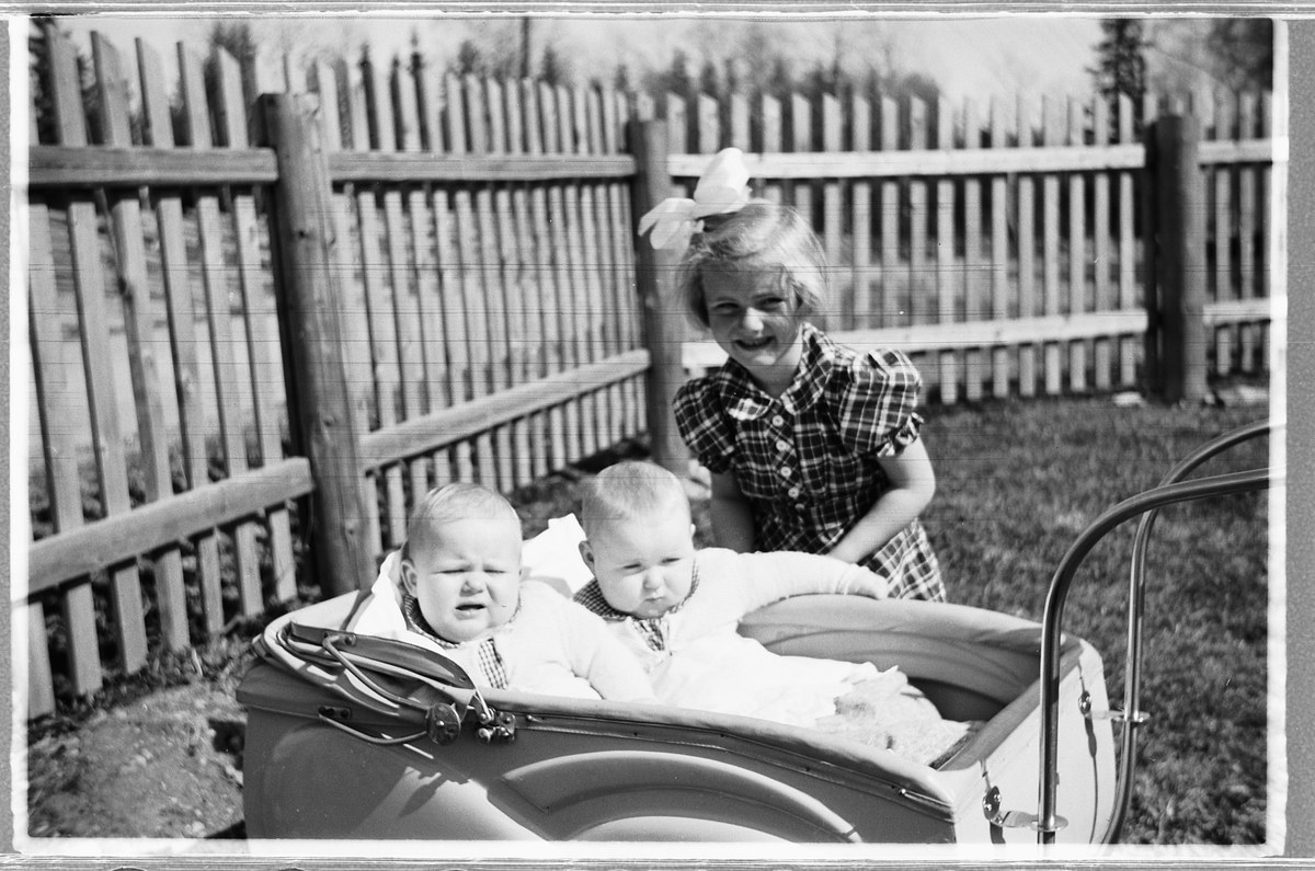 Drei Kinder in einem Auto, mit einem lachenden Mädchen im Vordergrund, einem Zaun und Bäumen im Hintergrund und grasbewachsenem Boden.