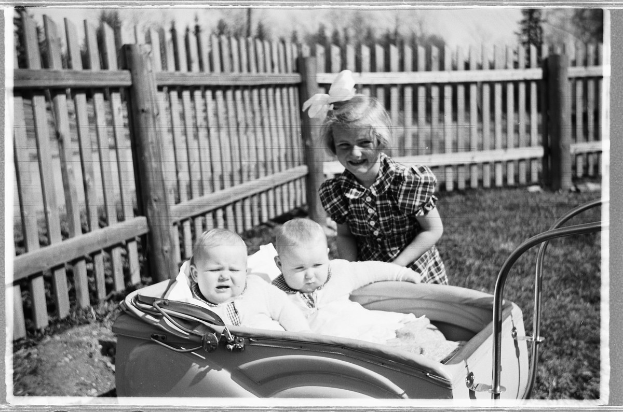Drei Kinder in einem Auto, mit einem lachenden Mädchen im Vordergrund, einem Zaun und Bäumen im Hintergrund und grasbewachsenem Boden.