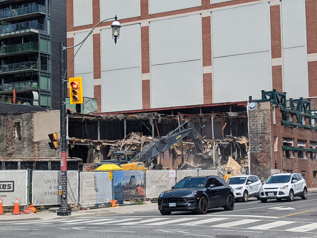 Ein abgerissenes Gebäude in der Mitte einer Straße, mit Fahrzeugen, Absperrgittern, einem Verkehrsampelmast und einem Kran im Vordergrund, umgeben von Gebäuden mit Fenstern und Geländern, unter einem sichtbaren Himmel.