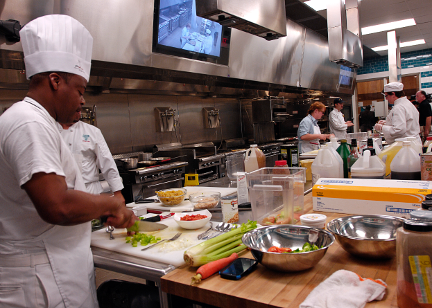 Eine Gruppe von Köchen in einer Küche beim Zubereiten von Speisen auf einem Schneidbrett, wobei ein Koch im Vordergrund in einer weißen Schürze und einer weißen Haube steht und ein Messer hält und von Utensilien, Schüsseln, Gemüse, einem Mobiltelefon, Flaschen und anderen Gegenständen umgeben ist, mit einem Fernseher und Deckenleuchten im Hintergrund.
