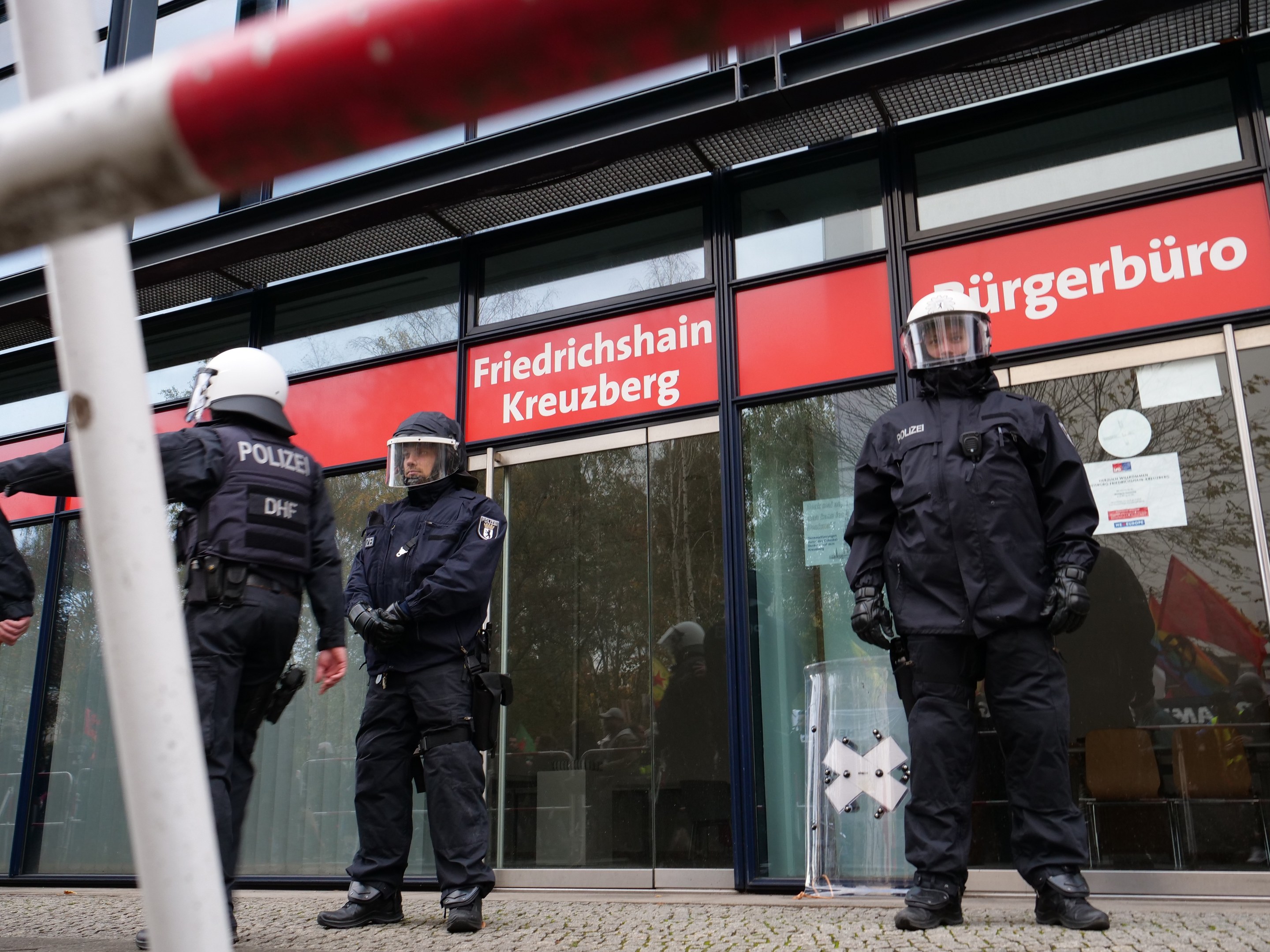 Polizeibeamte in Uniform vor einem Glasfrontgebäude mit einer Stange links und Schildern mit "Friedrichshain Kreuzberg"-Text im Hintergrund.
