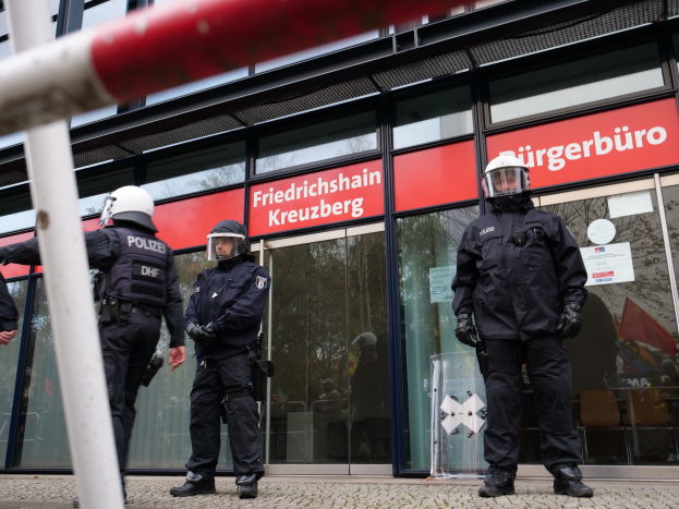 Polizeibeamte in Uniform vor einem Glasfrontgebäude mit einer Stange links und Schildern mit "Friedrichshain Kreuzberg"-Text im Hintergrund.